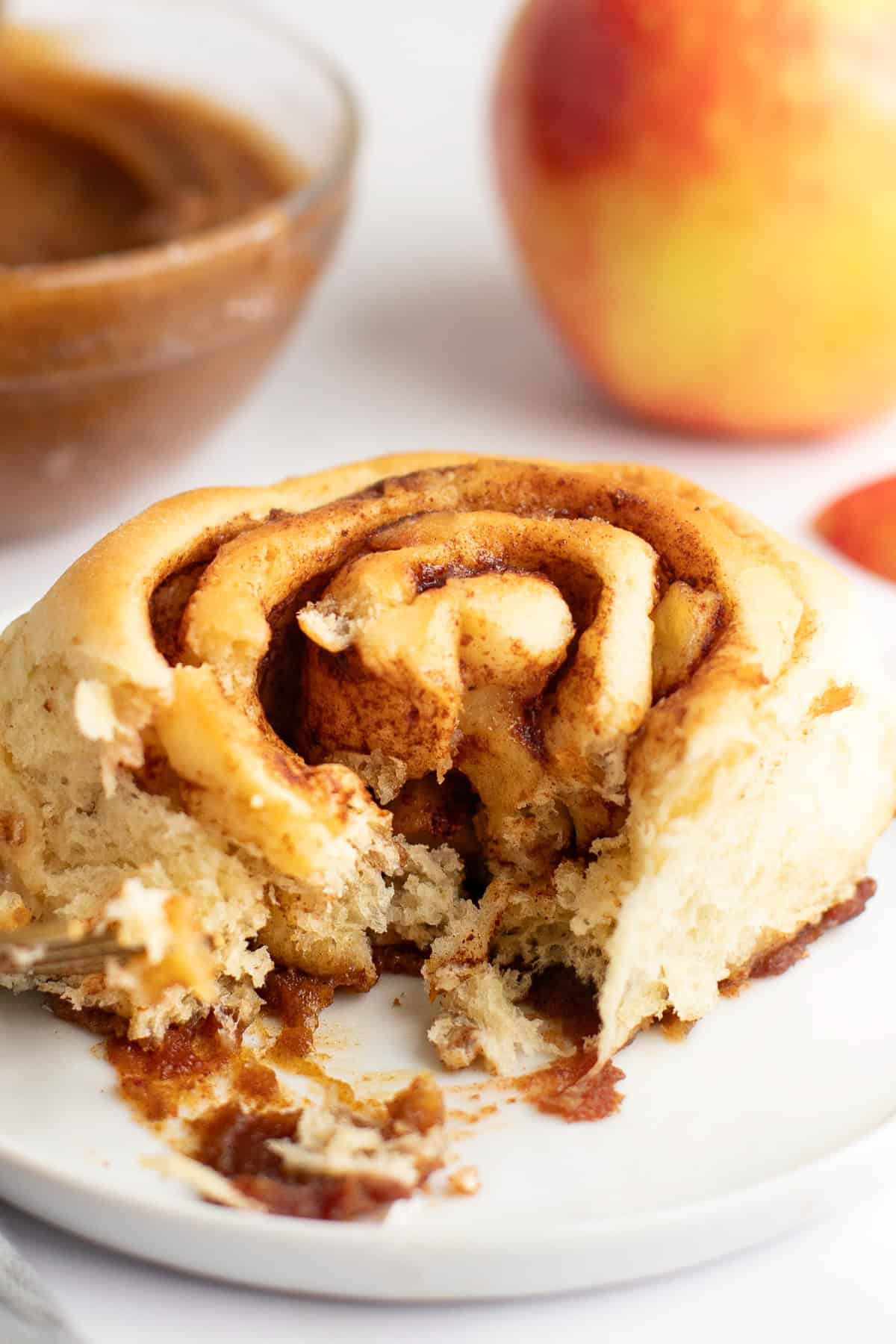 A partially eaten cinnamon roll on a white plate with a bowl of filling and an apple in the background.