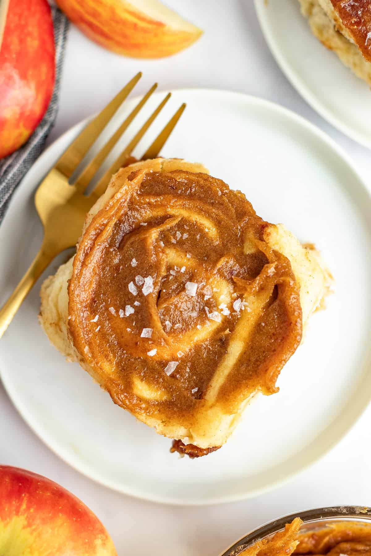 A single cinnamon roll with frosting and a sprinkle of sea salt is on a white plate with a gold fork. Slices of a red apple are partially visible in the background.