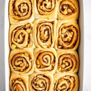 A rectangular baking dish filled with twelve freshly baked cinnamon rolls, each with a visible swirl pattern, on a white surface.