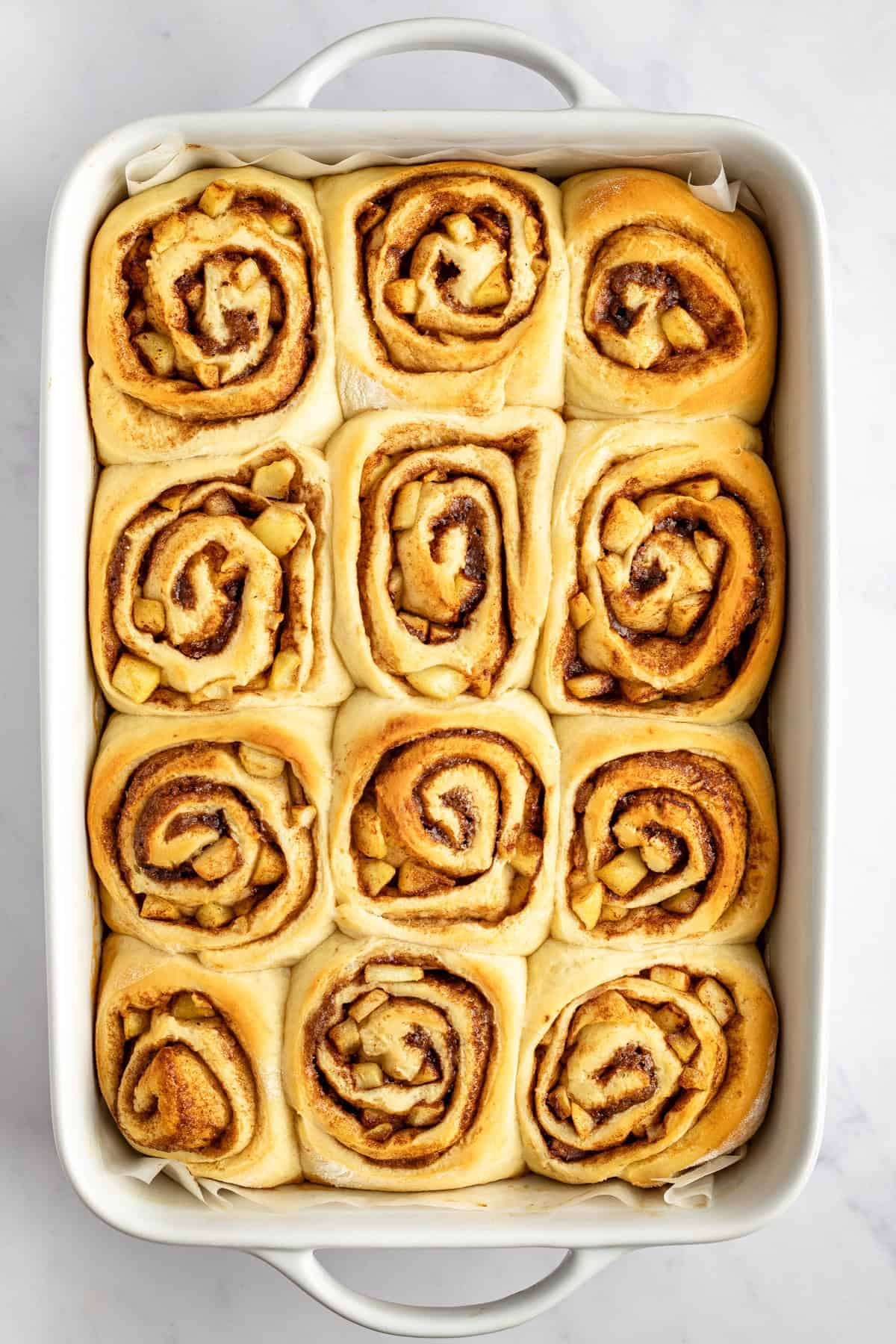 A rectangular baking dish filled with twelve freshly baked cinnamon rolls, each with a visible swirl pattern, on a white surface.