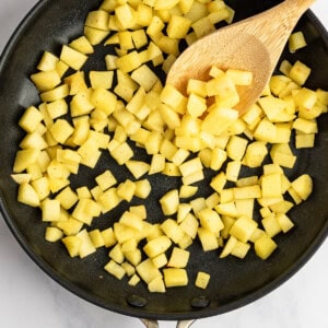 Diced potatoes cooking in a black skillet with a wooden spoon on a white marble surface.