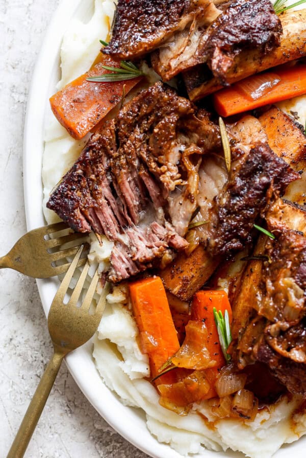 A plate of tender braised beef short ribs with carrots, garnished with rosemary, served on a bed of creamy mashed potatoes. Two forks are placed beside the dish.