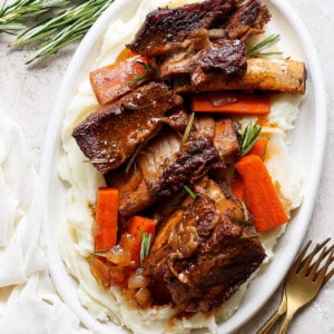 A white oval plate with mashed potatoes, roasted carrot slices, and beef short ribs garnished with fresh rosemary sprigs. A fork and knife are placed beside the plate on a white surface.