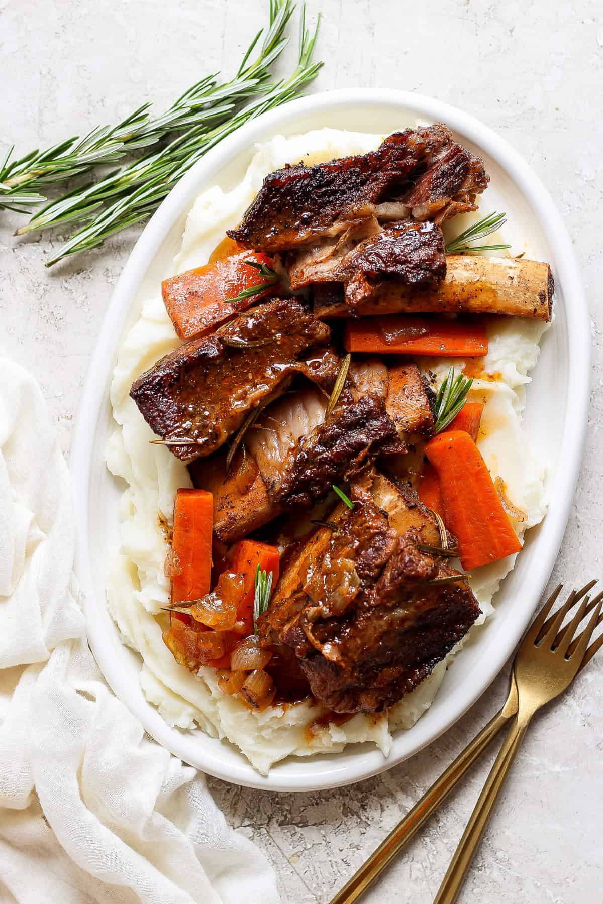 A white oval plate with mashed potatoes, roasted carrot slices, and beef short ribs garnished with fresh rosemary sprigs. A fork and knife are placed beside the plate on a white surface.