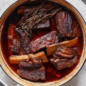 A large pot filled with braised short ribs in a rich, dark sauce, garnished with herbs on a light gray surface.