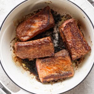 Four browned meat pieces are in a white, enameled Dutch oven on a light surface.