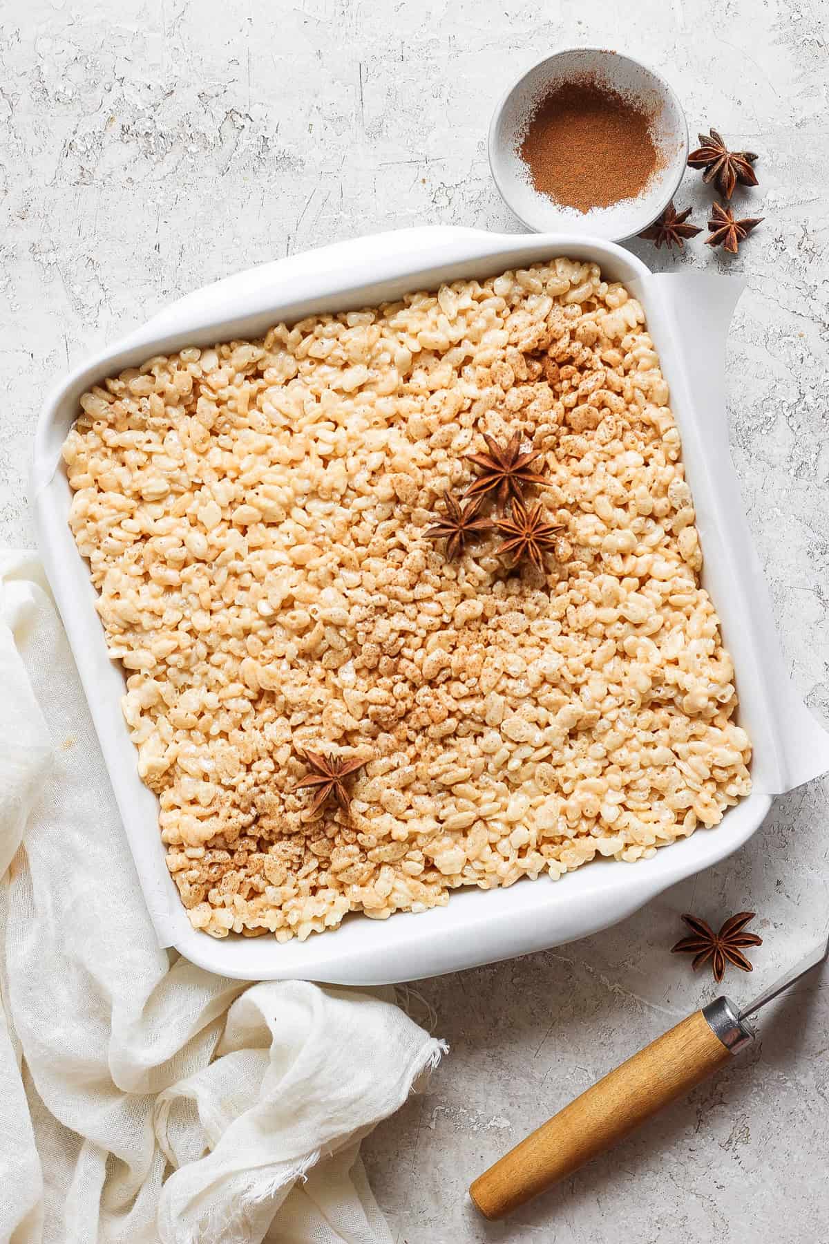 A square white baking dish filled with rice crispy treats, garnished with star anise and cinnamon powder in the top right corner. A small bowl with cinnamon powder is beside the dish on the counter.