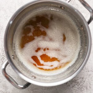 A metal pot with handles containing a frothy liquid that appears to be browning. The pot is placed on a light, textured surface.