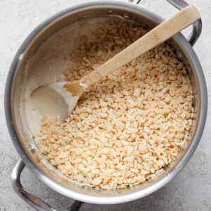 A pot containing a mixture of melted ingredients and rice cereal being stirred with a wooden spoon on a gray surface.