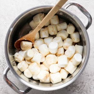 A pot filled with large marshmallows and a wooden spoon placed on a textured surface.