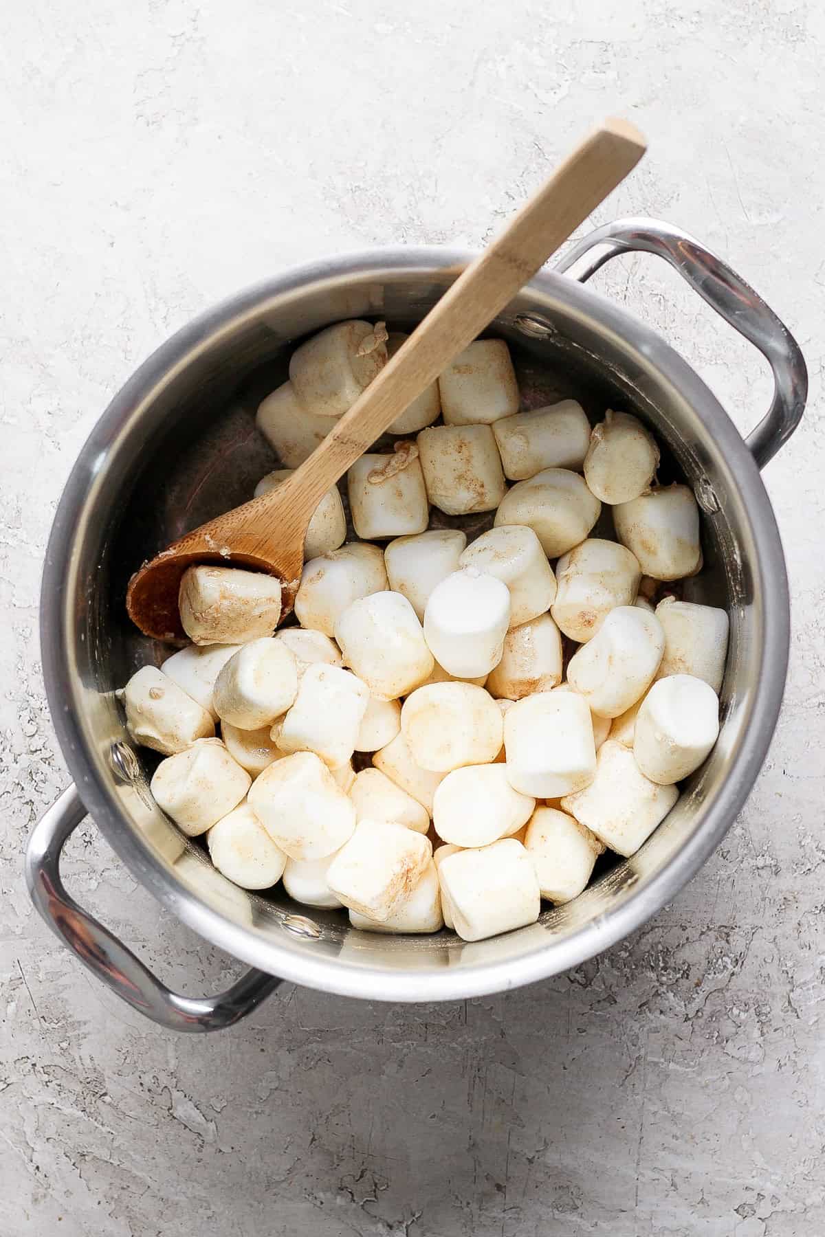 A pot filled with large marshmallows and a wooden spoon placed on a textured surface.