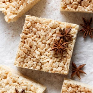 A square rice crispy treat topped with star anise spices, placed on a light-colored surface. Other identical treats and star anise pieces are shown around it.