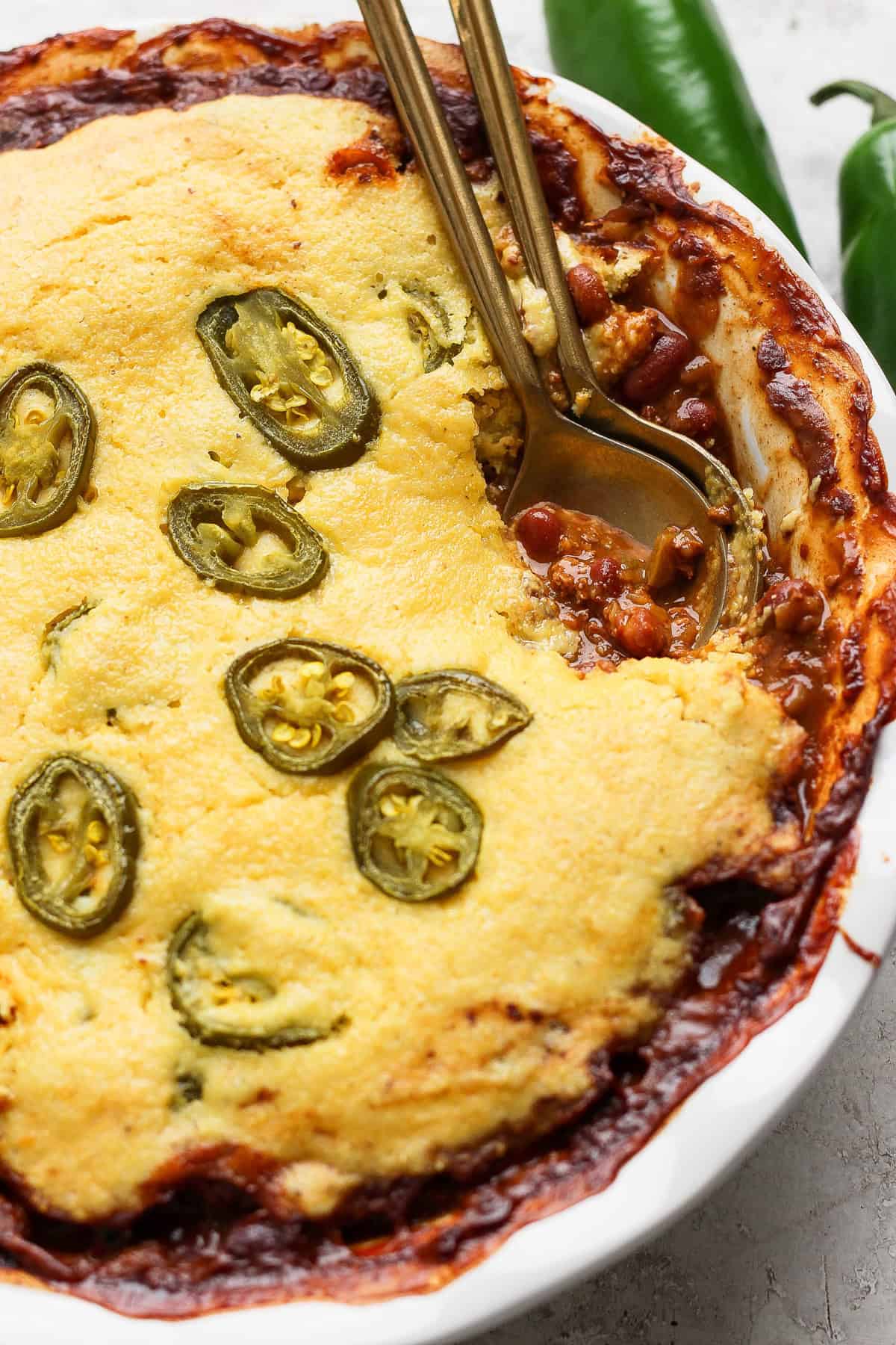 A baked dish in a white pie dish features a golden cornbread topping with sliced jalapeños, partially revealing a chili filling underneath. Two metal spoons are placed in the dish.