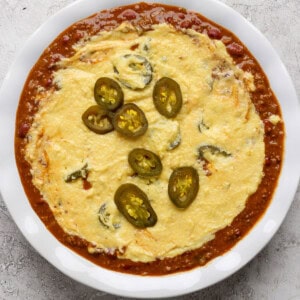 A white dish filled with melted cheese topped with sliced jalapeños, surrounded by a red tomato-based sauce.