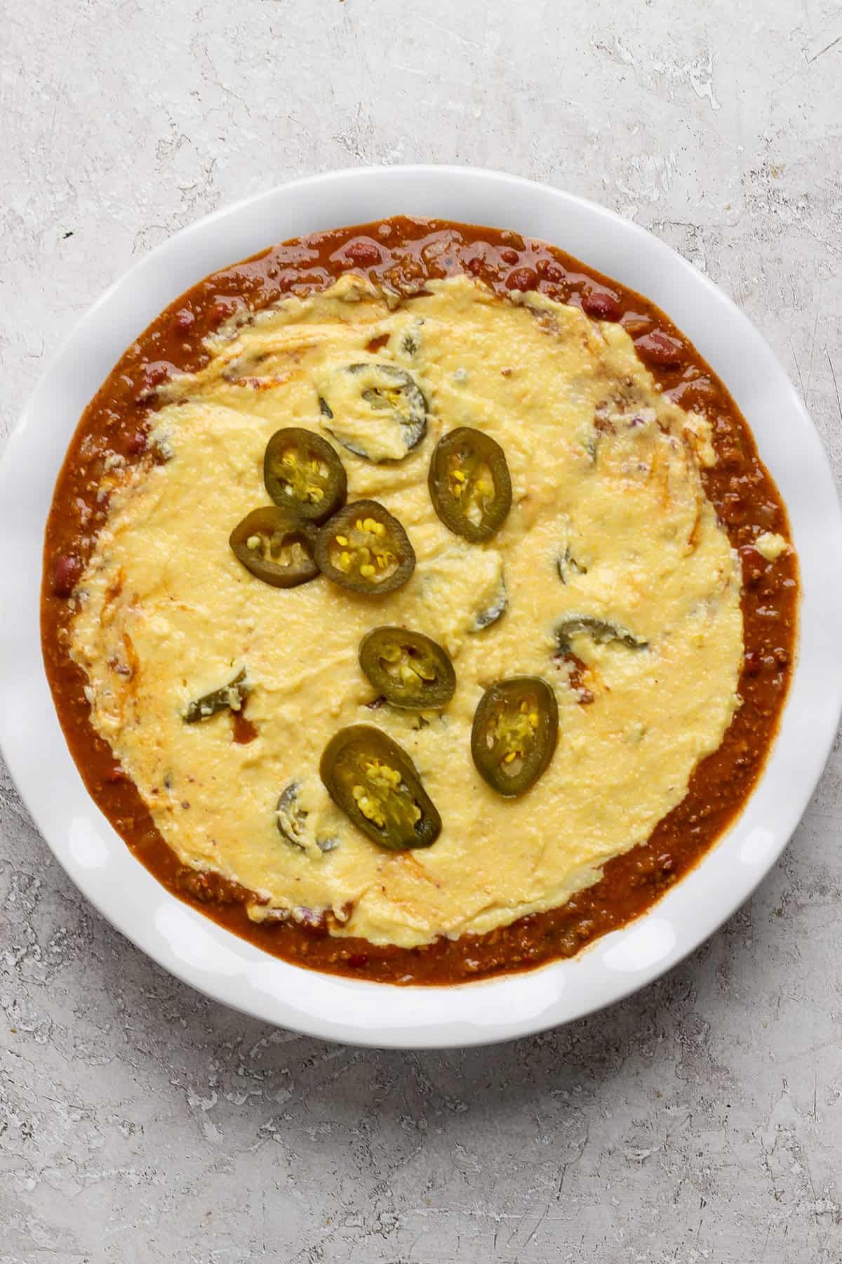 A white dish filled with melted cheese topped with sliced jalapeños, surrounded by a red tomato-based sauce.