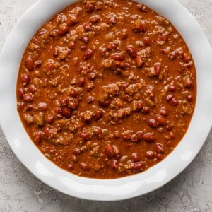 A white bowl filled with chili, containing ground meat, beans, and a thick tomato-based sauce, placed on a textured gray surface.