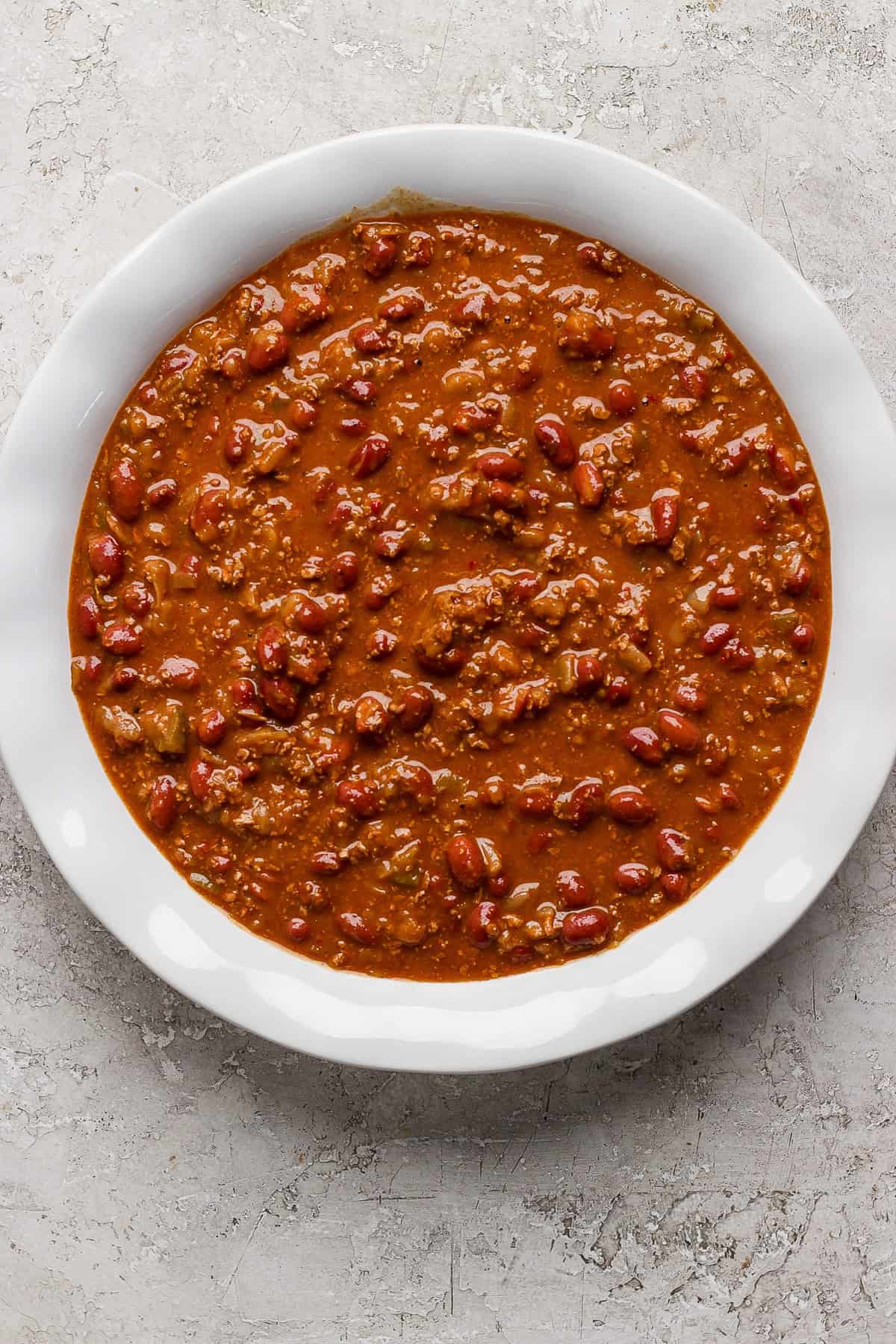 A white bowl filled with chili, containing ground meat, beans, and a thick tomato-based sauce, placed on a textured gray surface.