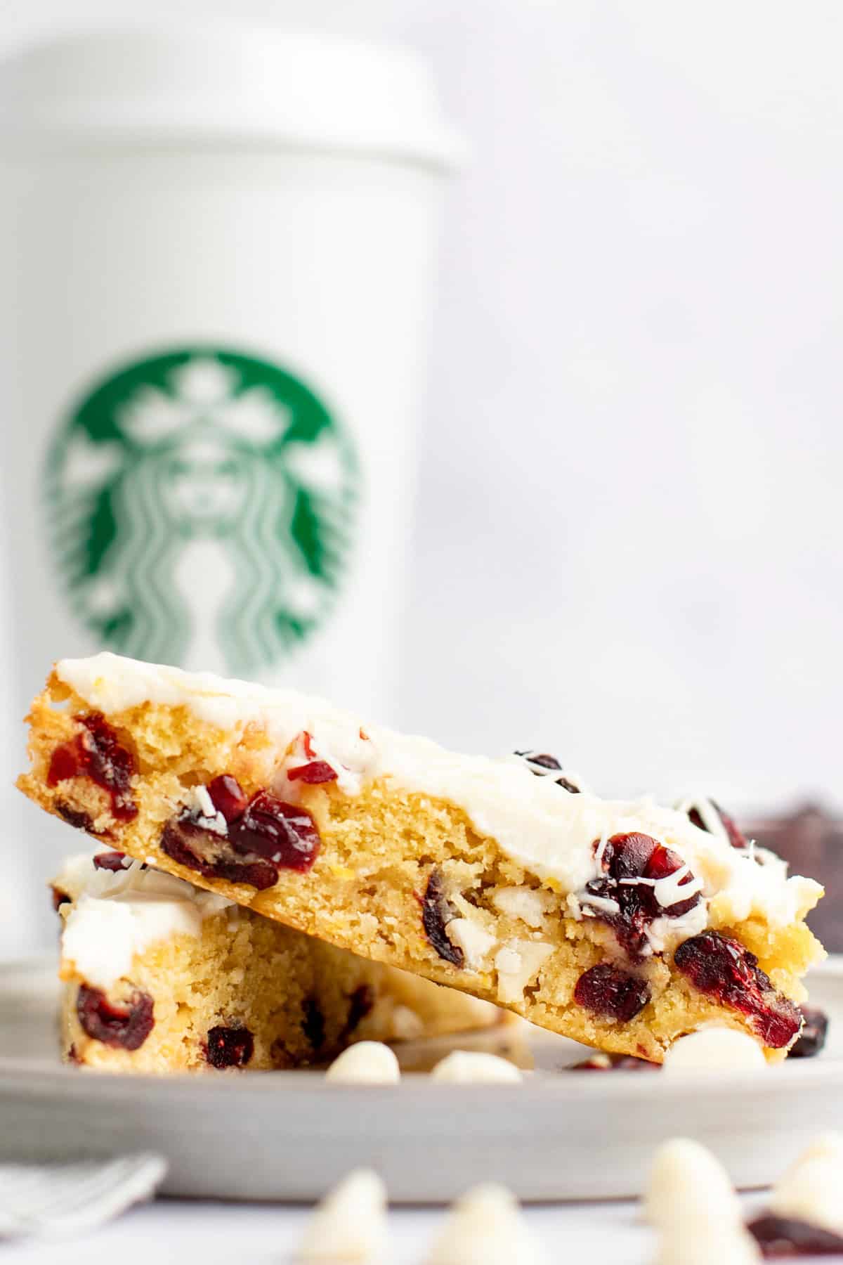 Two triangular slices of a white chocolate cranberry bar are stacked on a plate in front of a white Starbucks coffee cup.