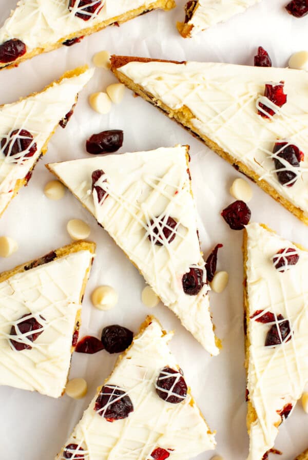 Triangular slices of a white chocolate and cranberry dessert bar are arranged on a white background, garnished with dried cranberries and white chocolate drizzle. White chocolate chips are scattered around.