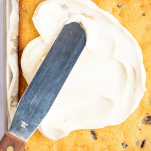 A metal spatula is spreading white frosting on a rectangular, baked cake in a square pan. The cake has visible chocolate chips.