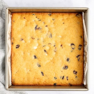 A square baking pan with a freshly baked golden-brown cake featuring scattered dark and light spots, rests on a marble surface beside a striped fabric.