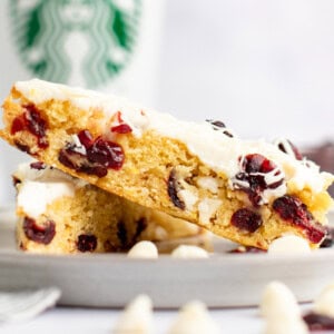 A close-up of two white chocolate cranberry bars stacked on a plate with a Starbucks cup in the background. The bars have a creamy white frosting and visible dried cranberries.