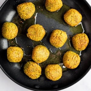 A skillet with twelve golden-brown falafel balls frying in oil on a light-colored surface.