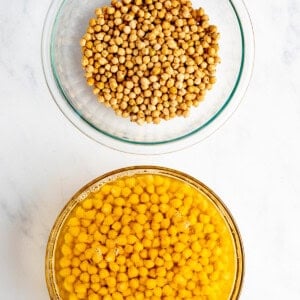 Two glass bowls on a marble surface: one with dried chickpeas and the other with soaked chickpeas in water.