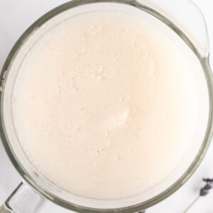 A clear glass bowl filled with a white, granular substance, viewed from above. Two sprigs of lavender are placed beside the bowl on a white surface.