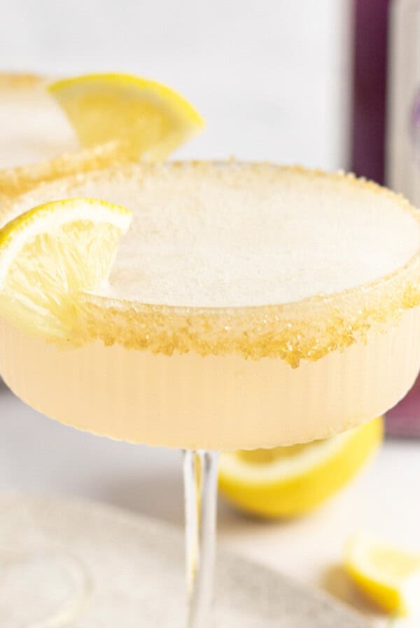 Two cocktails with frothy tops in coup glasses, each garnished with a lemon wedge and a sugar-rimmed edge. A bottle of purple liquid is partially visible in the background.