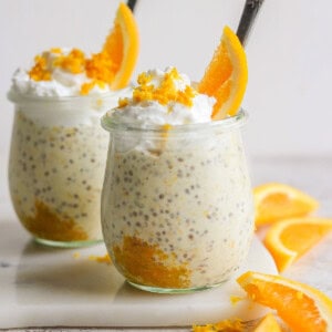 Two glass jars filled with creamy creamsicle overnight oats, topped with whipped cream and orange slices, arranged on a white surface with additional orange wedges nearby.