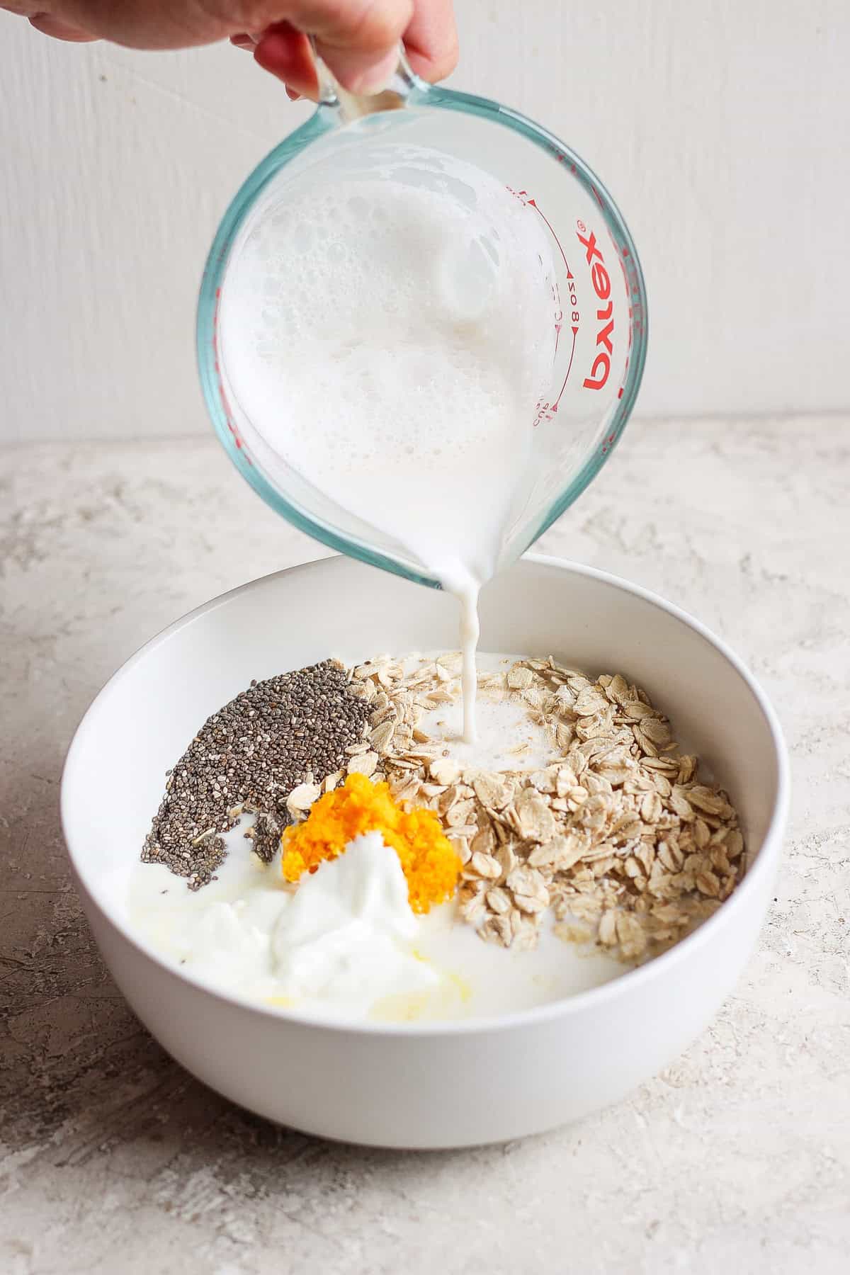 A hand pours milk from a measuring cup into a white bowl containing oats, chia seeds, orange zest, and yogurt, creating the perfect base for creamsicle overnight oats.