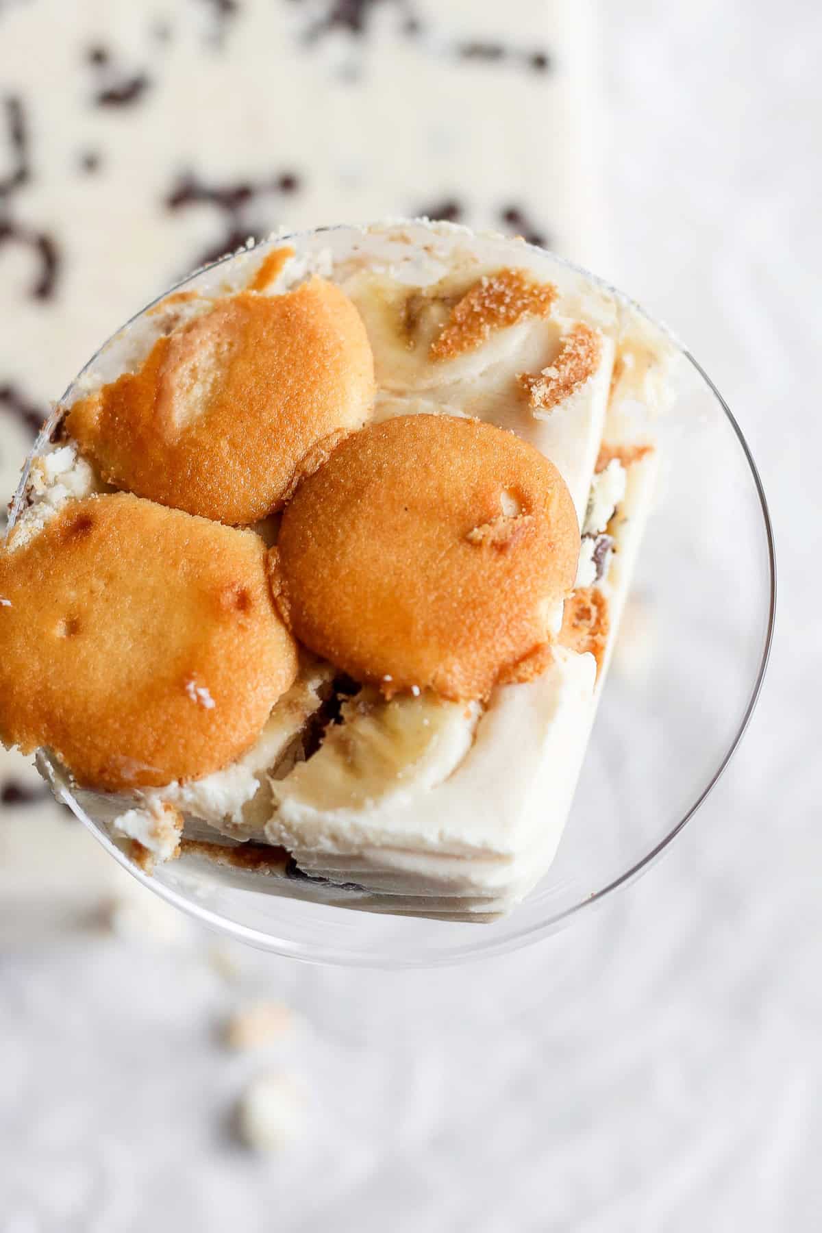 A glass bowl filled with a dessert made of layered vanilla wafers, slices of banana, and creamy pudding. The dessert, reminiscent of a peanut butter banana ice box cake, is topped with vanilla wafers.