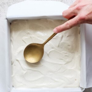 A hand holding a spoon smooths out a white batter for peanut butter banana ice box cake in a parchment-lined square baking pan on a textured countertop.