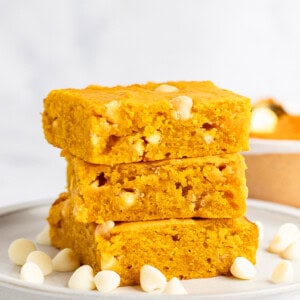 Three slices of golden yellow pumpkin blondies stacked on a plate, surrounded by white chocolate chips.