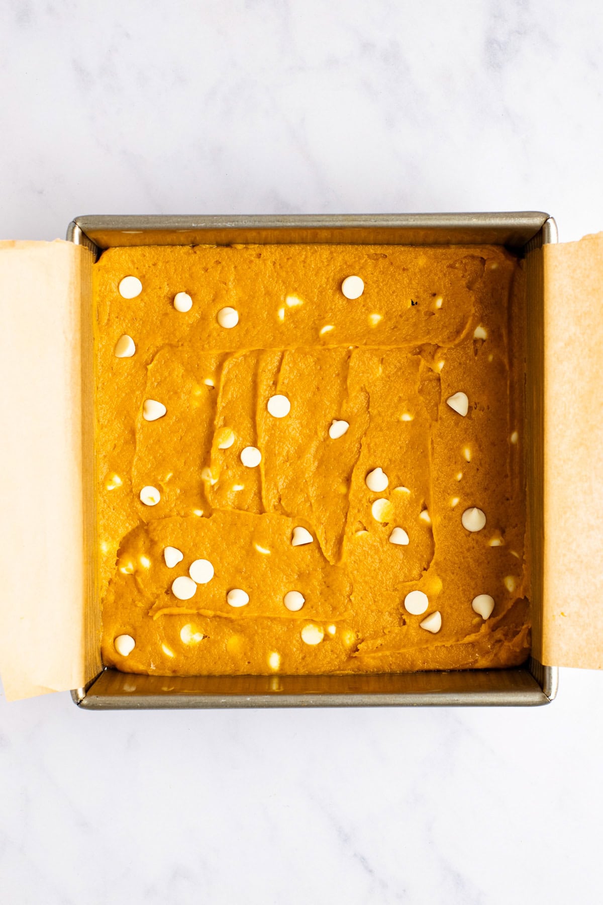 A square baking pan filled with unbaked batter topped with scattered white chocolate chips, lined with parchment paper on a marble countertop.