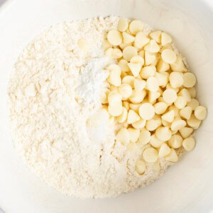 A white bowl containing a mixture of flour, baking powder, and white chocolate chips on a white background.