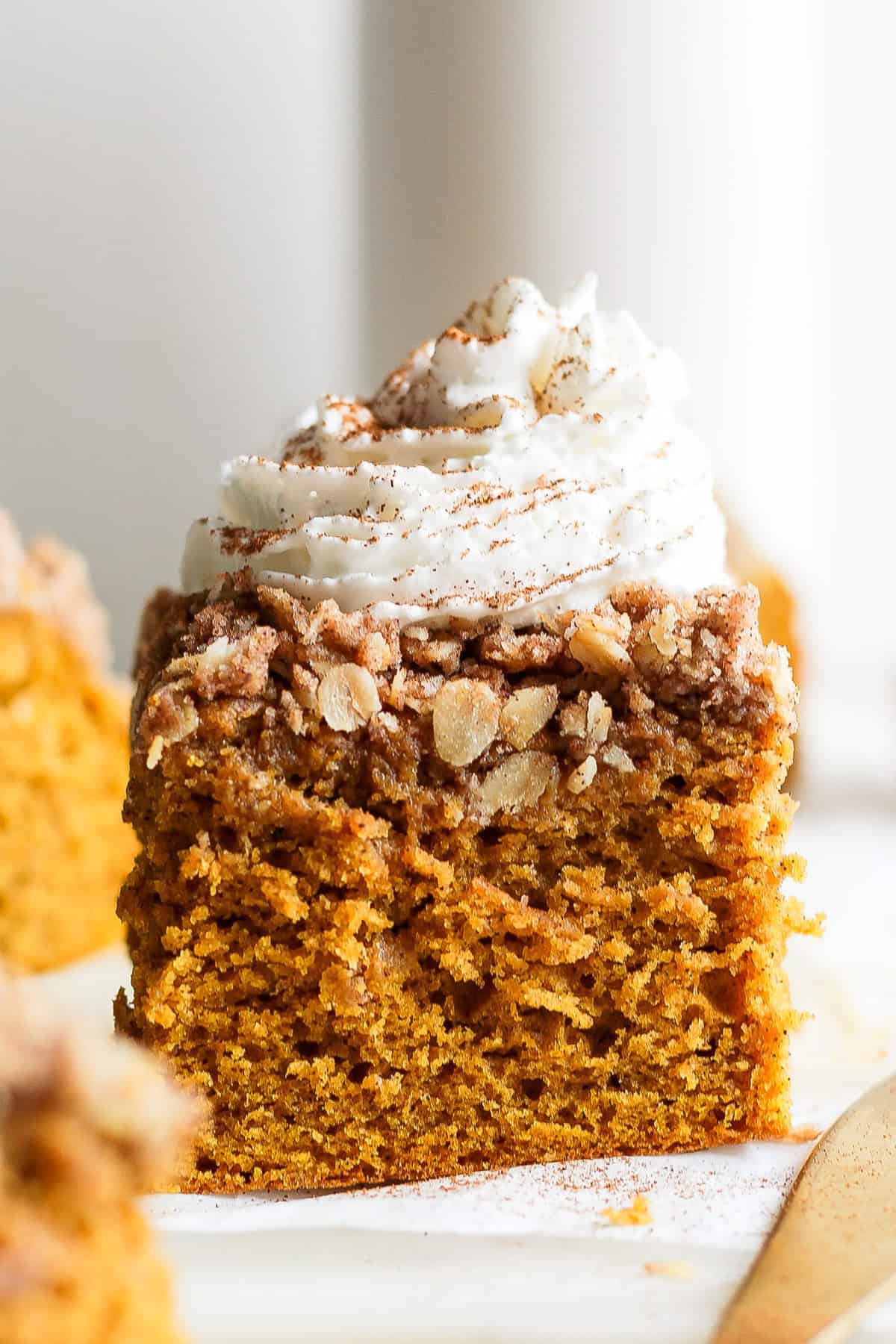 A slice of pumpkin coffee cake topped with whipped cream and a sprinkle of cinnamon on a white surface.