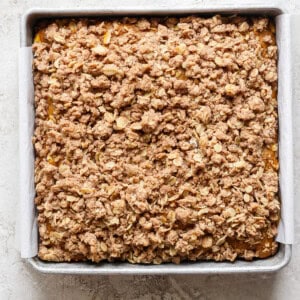 A square metal baking pan filled with a baked oatmeal fruit crumble topped with a crispy, golden-brown oat and cinnamon streusel. The pan is lined with parchment paper.