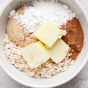 A white bowl containing oats, brown sugar, flour, cinnamon, and two slabs of butter on a light-colored surface.