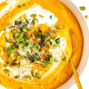 A bowl of creamy orange soup garnished with fresh microgreens, pumpkin seeds, black pepper, and a swirl of cream. A golden spoon rests inside the bowl.