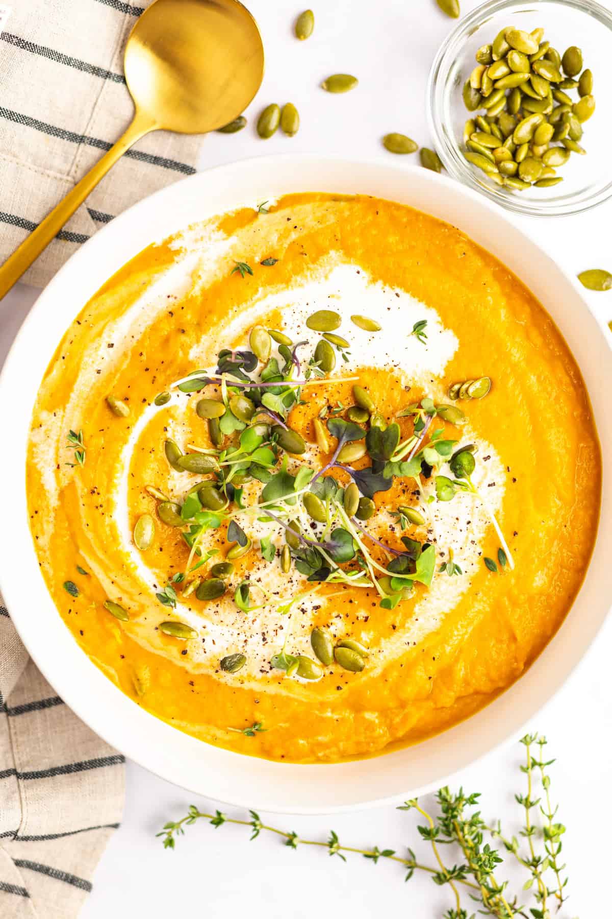 A bowl of creamy orange soup garnished with microgreens, pumpkin seeds, and a swirl of cream. A gold spoon, sprigs of fresh herbs, and a small bowl of pumpkin seeds are placed nearby.