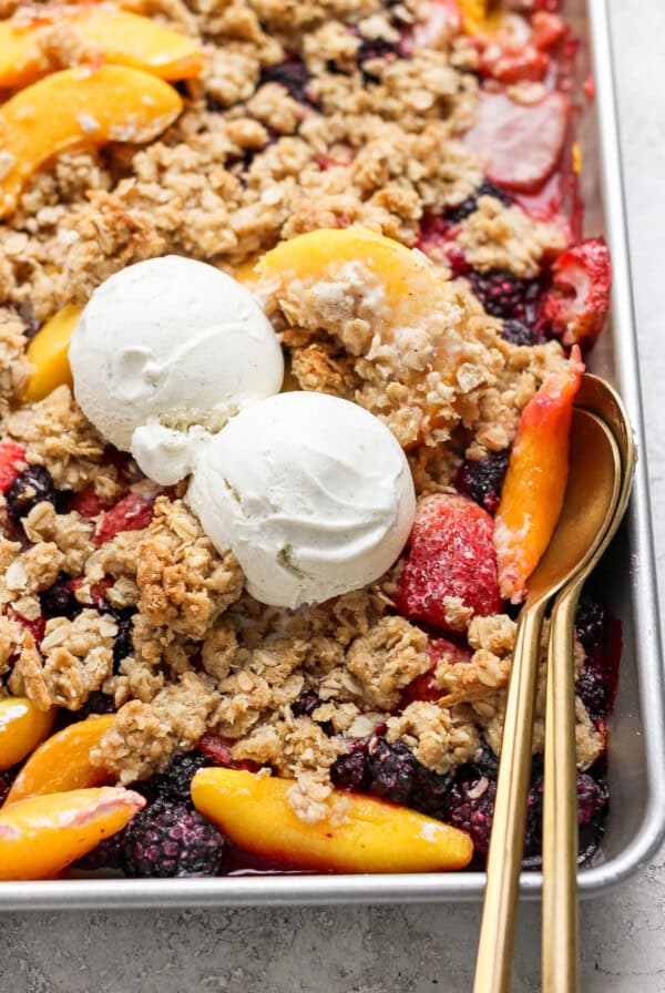 A baking dish with a mixed berry and peach crumble topped with two scoops of vanilla ice cream; a gold spoon rests on the right side of the dish.