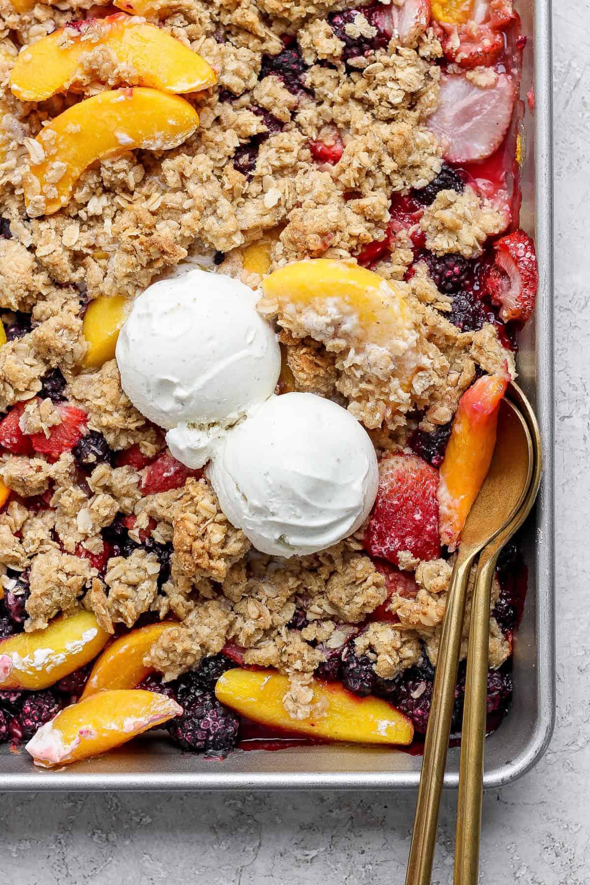 A tray of mixed fruit crumble topped with two scoops of vanilla ice cream and oat crumbles, with a serving spoon on the right side.