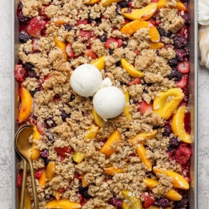 A rectangular baking tray filled with a mixed berry and peach crumble topped with two scoops of vanilla ice cream. A gold spoon rests on the left side of the tray.