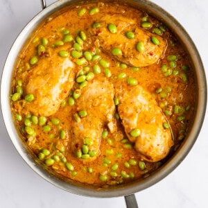 A stainless steel pan contains cooked chicken breasts in a golden sauce with green edamame.