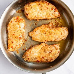 Four seasoned chicken breasts are cooking in a stainless steel skillet.