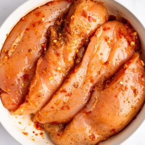 A bowl containing raw chicken breasts marinated with red pepper flakes and other spices.