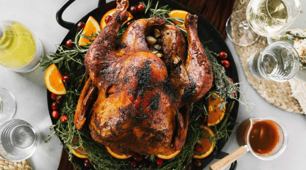 A roasted turkey garnished with herbs and orange slices is served on a black platter, accompanied by glasses of white wine, water, and a bowl of gravy.