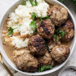 A plate of meatballs covered in gravy, served with mashed potatoes and garnished with fresh parsley. A fork and a small bowl of pepper sit adjacent to the plate.